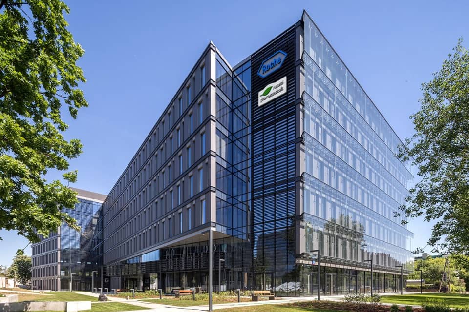 Natural Pharmaceuticals „Movisan® Collagen II” Modernus biurų pastatas su stikliniu fasadu šviesiu, saulėtu dieną.