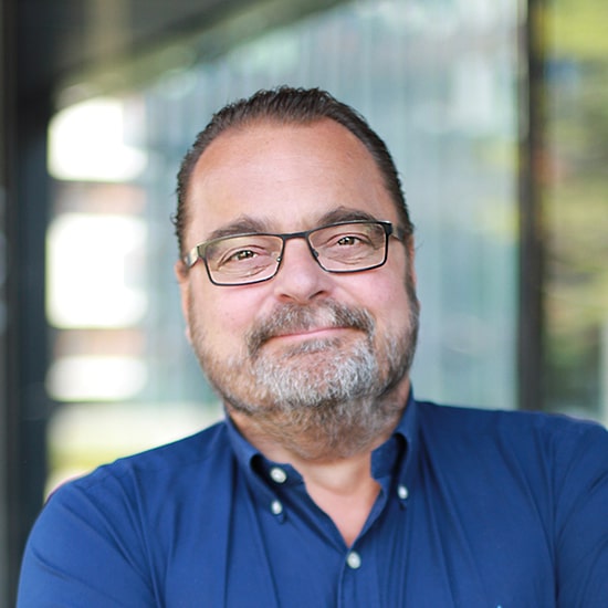 Natural Pharmaceuticals „Movisan® Collagen II” Vyras su akiniais ir mėlyna palaidine šypsosi, stovėdamas priešais langus biuro aplinkoje.