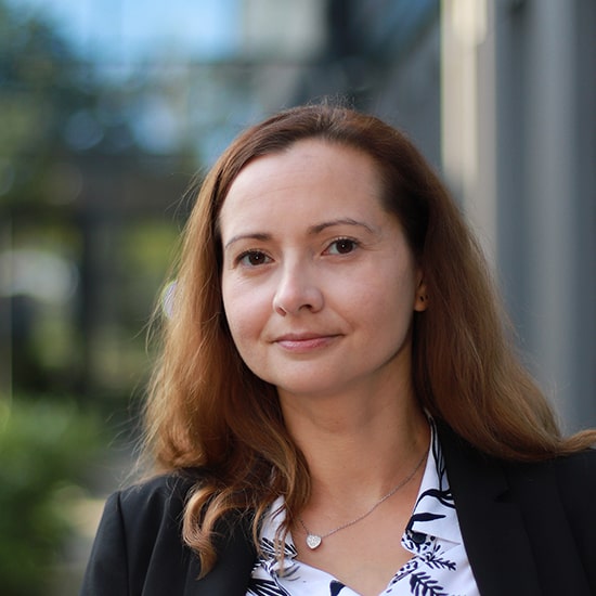 Natural Pharmaceuticals „Movisan® Collagen II” Vidutinio amžiaus moteris su šviesiais plaukais, dėvinti juodą švarką ir baltą marškinių su augalų raštu fone.