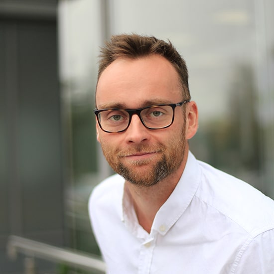 Natural Pharmaceuticals „Movisan® Collagen II” Vidutinio amžiaus vyras su akiniais ir baltu marškinėliu, šypsosi prie stiklinio pastato fasado.