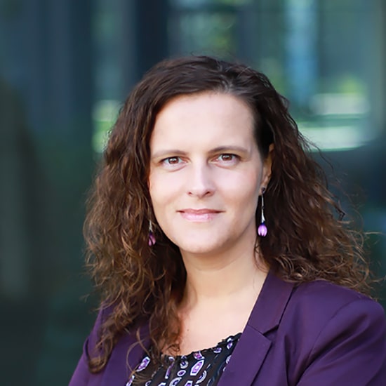 Natural Pharmaceuticals „Movisan® Collagen II” Vidutinio amžiaus moteris su garbanotais rudais plaukais, vilkinti violetinį švarką ir laikanti neutralų veido išraišką.