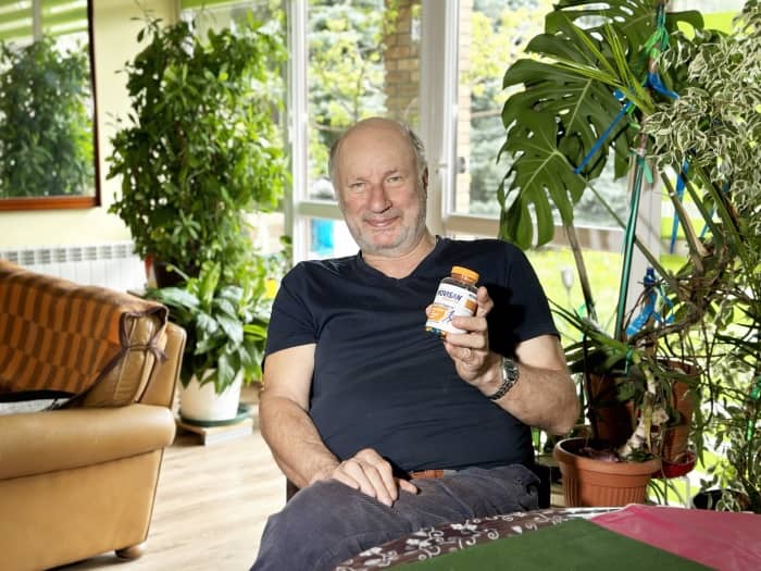 PNatural Pharmaceuticals „Movisan® Collagen II” Vyriškis kambaryje tarp daugybės žalumos sėdi ir laiko maisto papildų buteliuką.