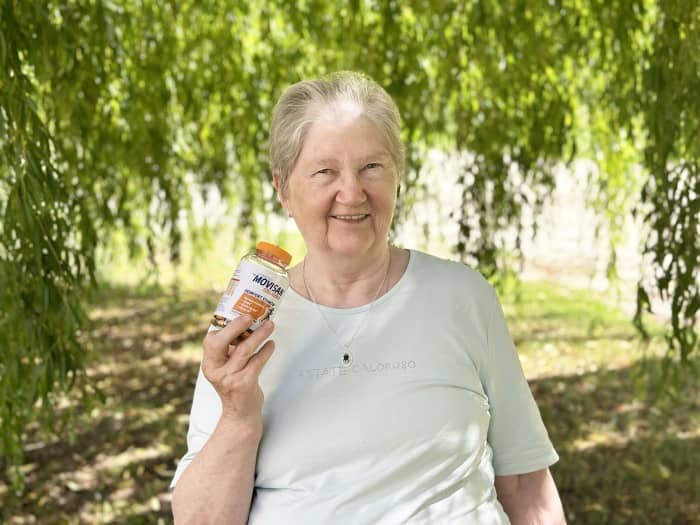 Natural Pharmaceuticals „Movisan® Collagen II” Vyresnės moters portretas gamtoje, laikantis Movisan maisto papildų buteliuką ir šypsantis.