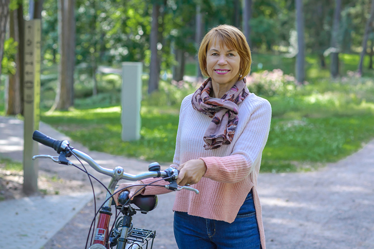 Natural Pharmaceuticals „Movisan® Collagen II” Vidutinio amžiaus moteris su skarele ir megztiniu stovi su dviračiu parke saulėtą dieną.